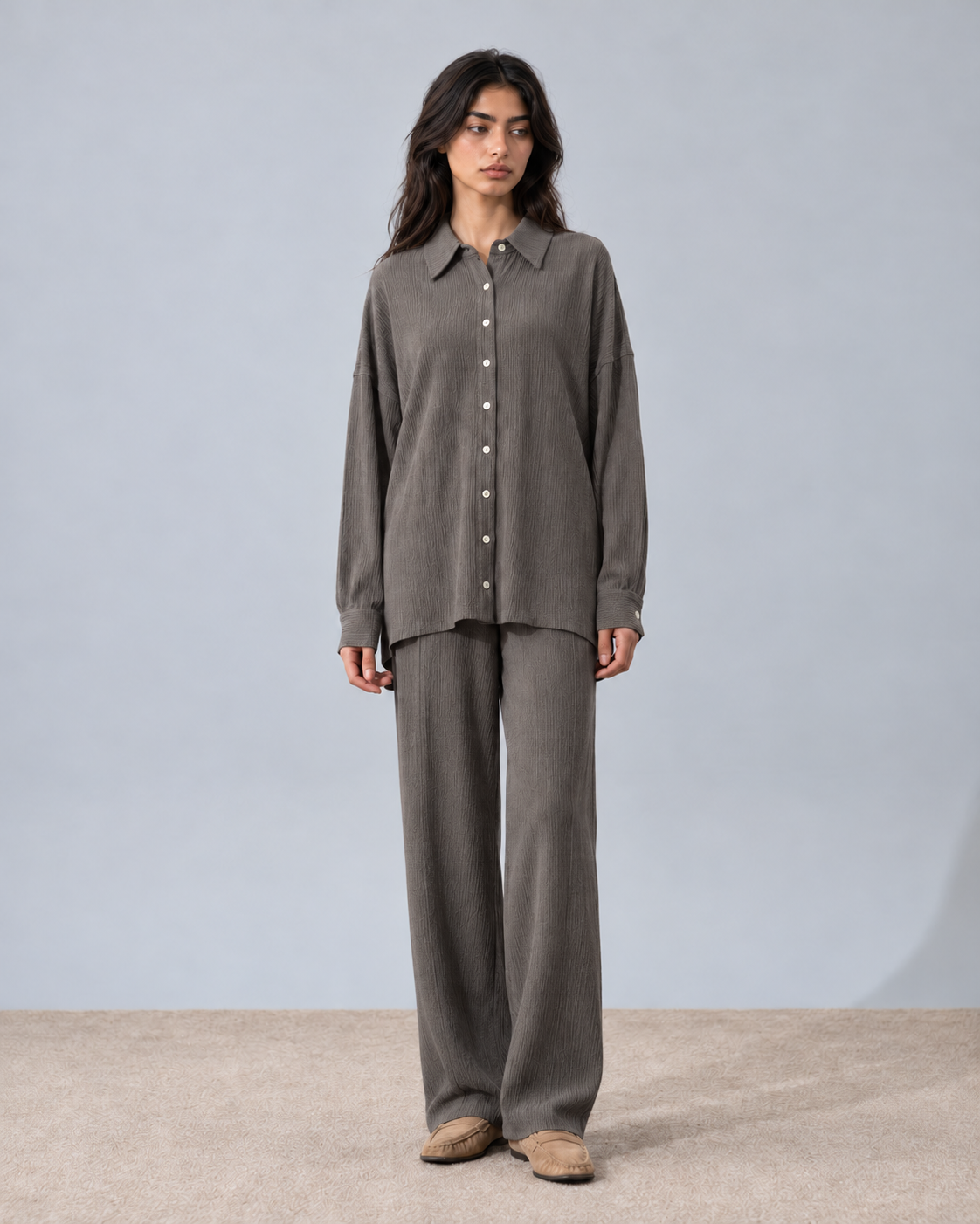 A woman stands against a light gray studio backdrop wearing a matching two-piece lounge set consisting of a long-sleeve button-front shirt and wide-leg pants in a taupe-gray color, paired with tan slip-on shoes.