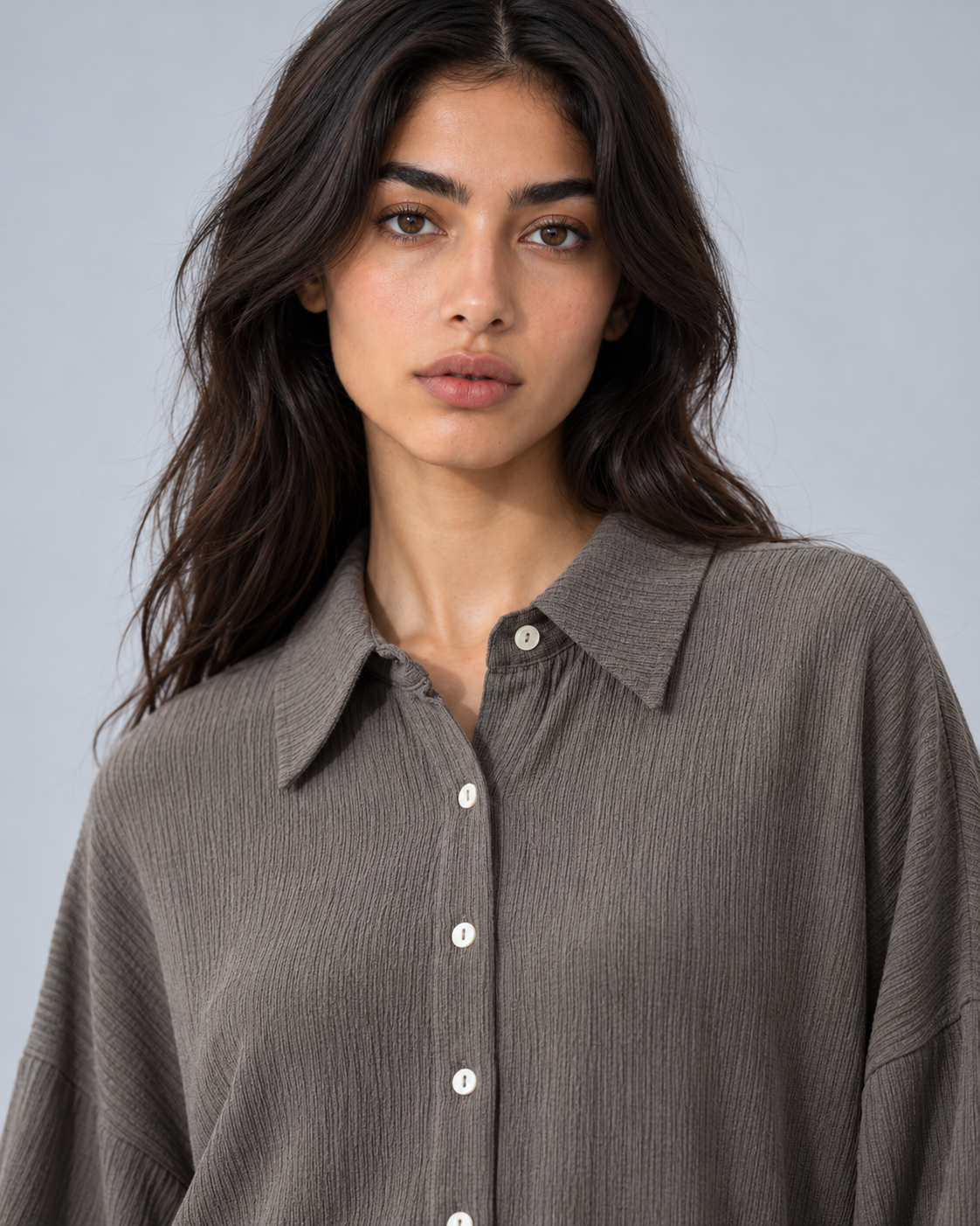 A woman wearing a textured, crinkled button-up shirt in a muted taupe-brown color, shown from the chest up against a solid light gray background.