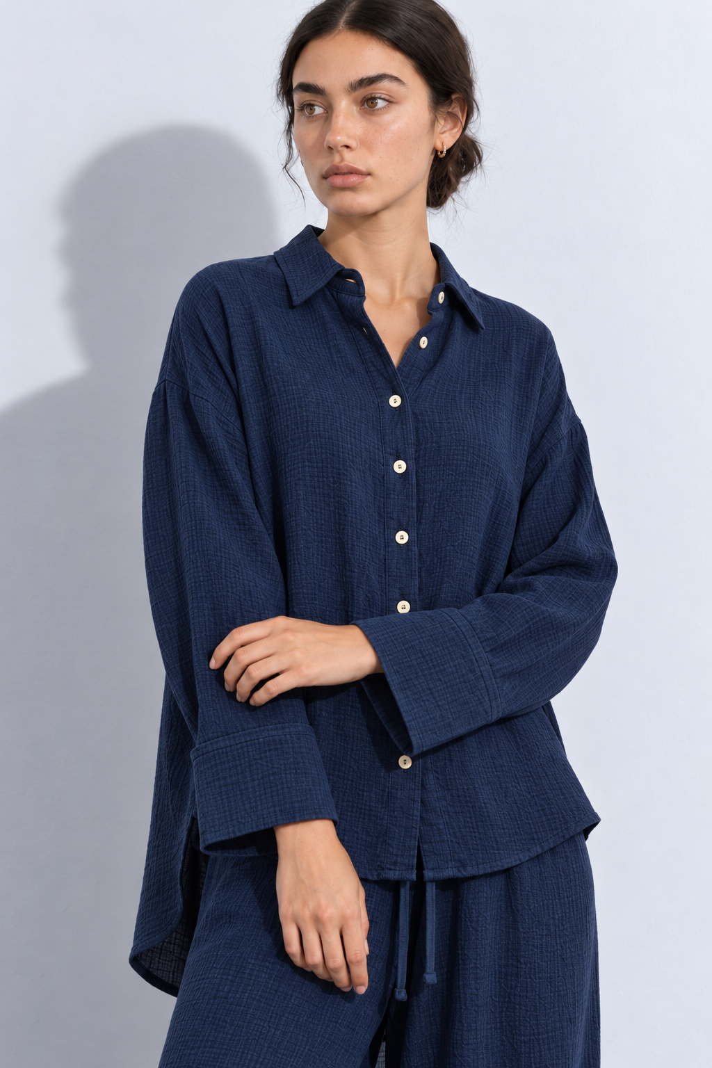 A woman wearing a dark navy textured button-up shirt and matching pants, standing against a light gray studio background with a soft shadow.